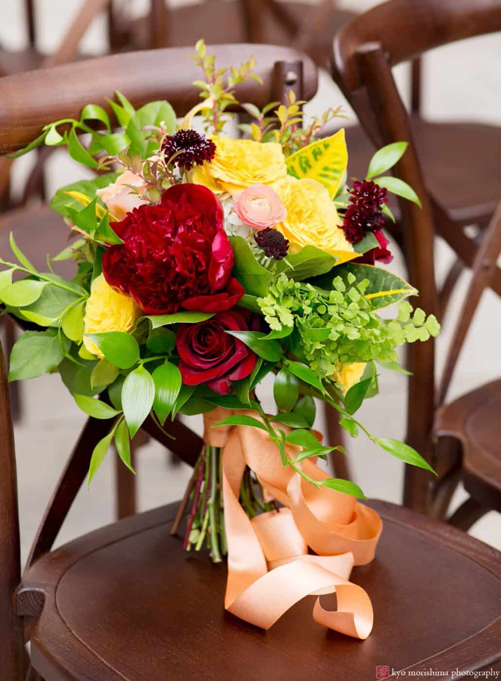 Colorful maroon and gold bouquet with dark red roses and peonies, yellow roses, pale blush ranunculus, ruby scabiosa, and croton petra leaves, tied with apricot ribbon. Wedding bouquet by Kristin Rockhill of Details of I Do, photographed by Kyo Morishima