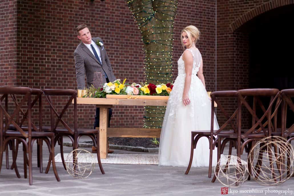 Bride (wearing BHLDN Ariane wedding gown) and groom stand at West Elm table decorated with maroon and gold flowers by Kristin Rockhill of Details of I Do, at 