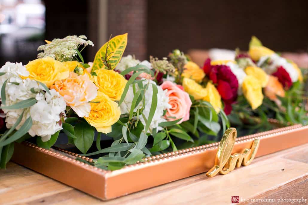 Detail of outdoor table decor with yellow, maroon, and blush roses and peonies and Croton leaf in a copper tray, styled by Kristin Rockhill of Details of I Do, photographed by Kyo Morishima