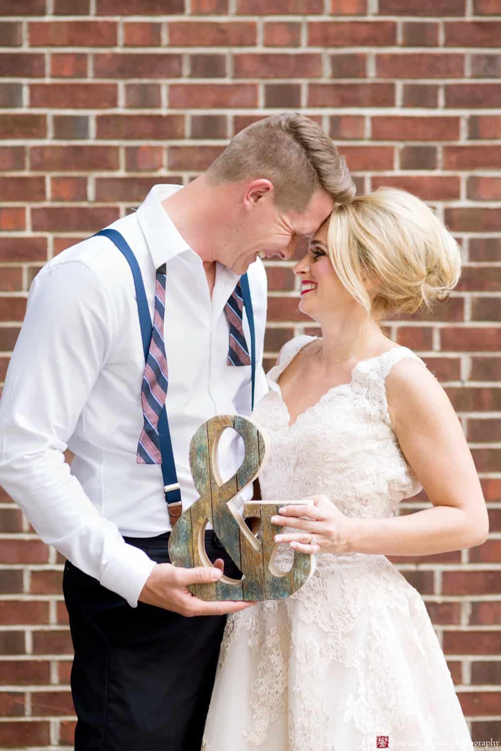 Bride and groom outside brick wall behind Nassau Inn in Princeton; bride wears BHLDN wedding dress and groom holds ampersand sign. Styled by Kristin Rockhill of Details of I Do, photographed by Kyo Morishima