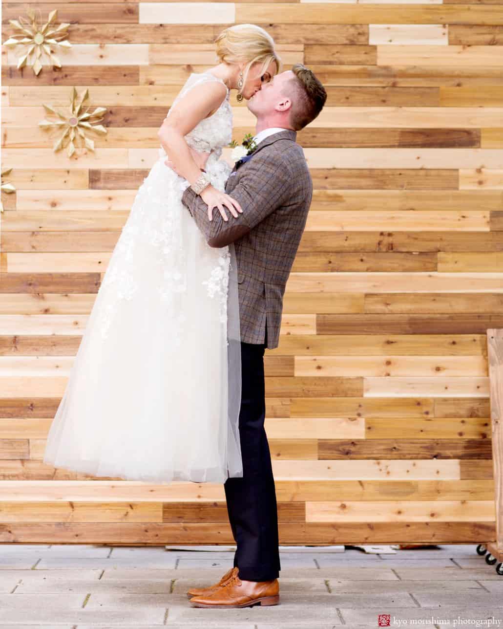 Bride wearing Ariane wedding gown from BHLDN kisses groom in front of wood backdrop at Nassau Inn 