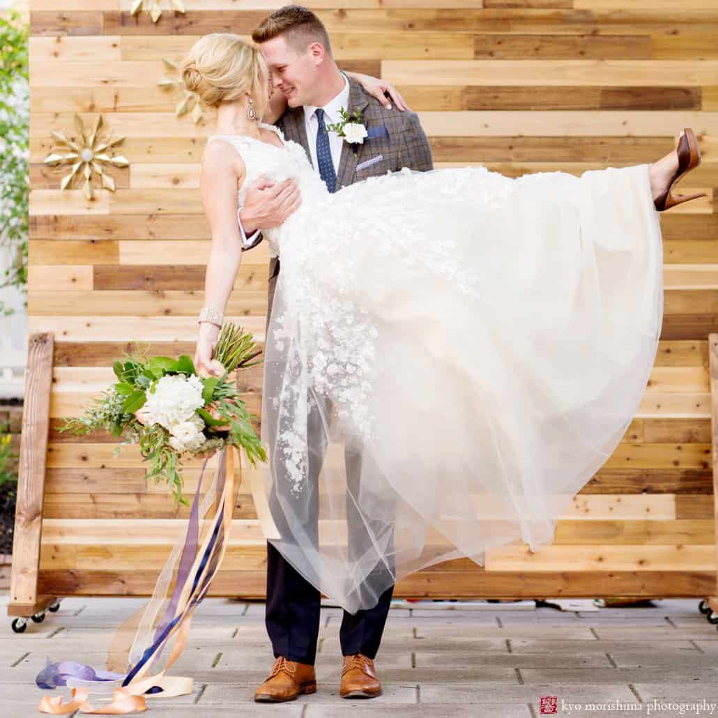 Groom holds bride wearing Ariane wedding gown from BHLDN in front of wood backdrop at Nassau Inn 