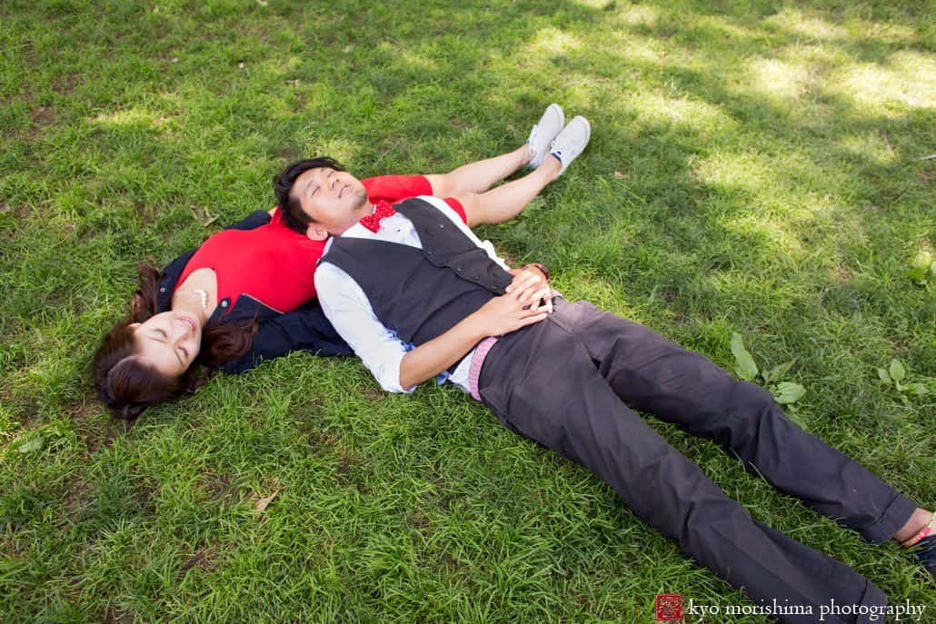 Woman wearing red dress lies on grass in Central Park with her fiancé resting on her; engagement photo photographed by Kyo Morishima