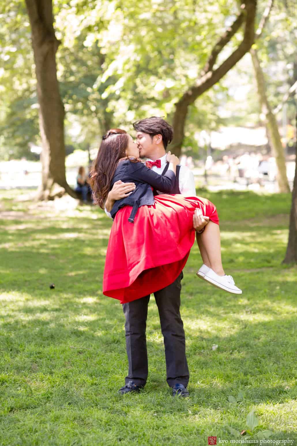 Man holds woman wearing red dress and gives her a kiss at the end of Central Park engagement photo session photographed by Kyo Morishima