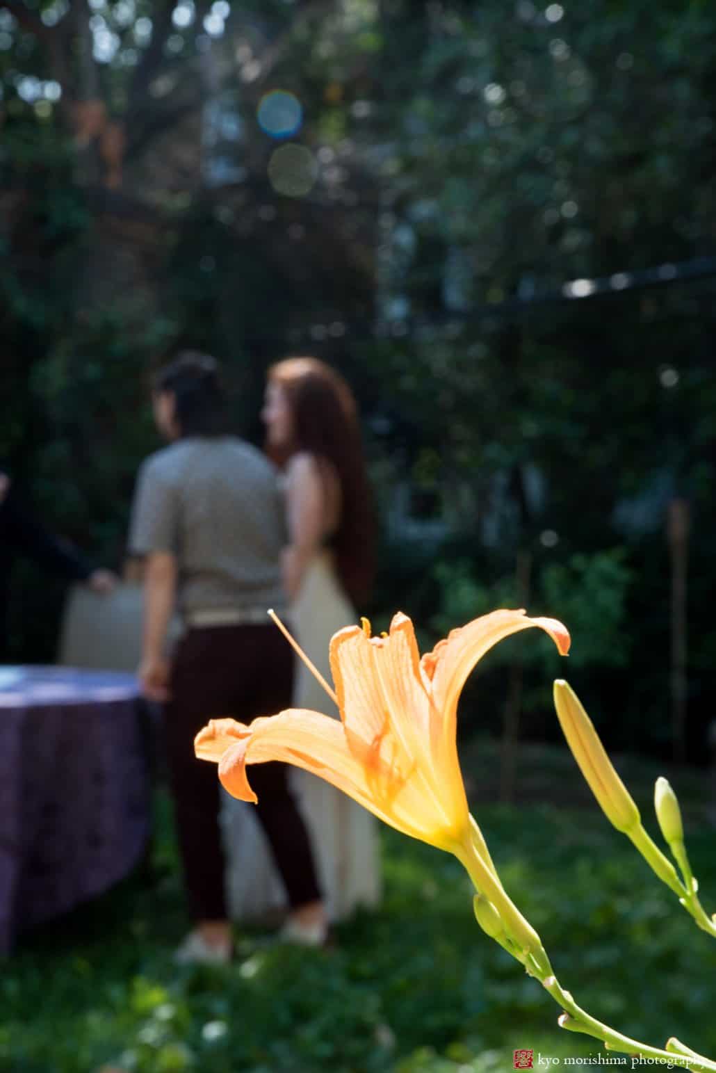 Daylily in the foreground as bride and her partner wait to sign the ketubah in backyard wedding ceremony, photographed by Kyo Morishima