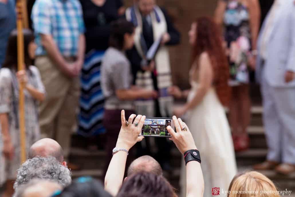 Guests snaps a photo with their phone during West Philadelphia outdoor wedding ceremony, photographed by Kyo Morishima