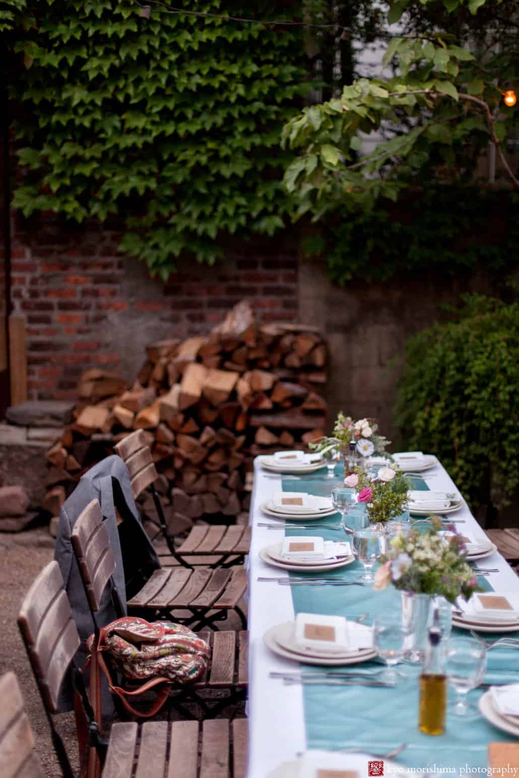 Long tables set with teal silk runners and centerpieces by Opalia Flowers with wood stack in the background; a Frankies 457 Spuntino wedding reception photographed by Kyo Morishima