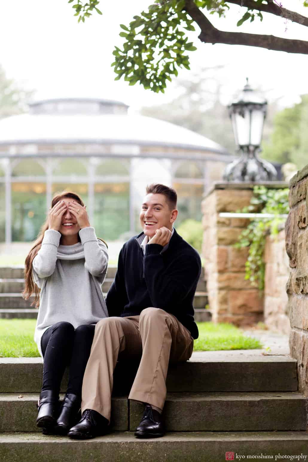 Unique engagement photo as woman covers her eyes and fiancé smiles at camera, photographed at Jasna Polana with glass ballroom in the background