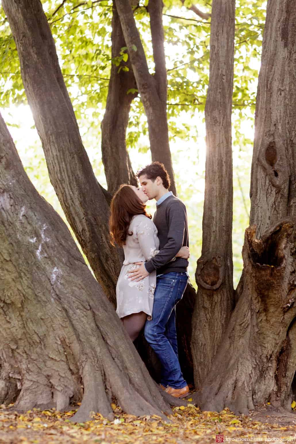 Prospect Park engagement photo: couple kisses while standing among tree trunks, photographed by Kyo Morishima