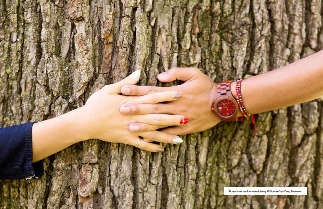 A couple clasp hands around a big oak tree trunk; the woman wears a vibrant gel manicure with a different finish on each fingernail.