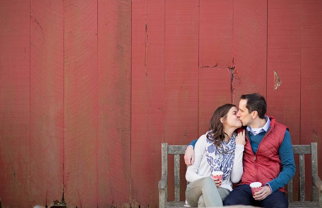 Candid engagement photographer in New Jersey: couple sit on a bench and kiss in front of a red barn at Terhune Orchards in Princeton