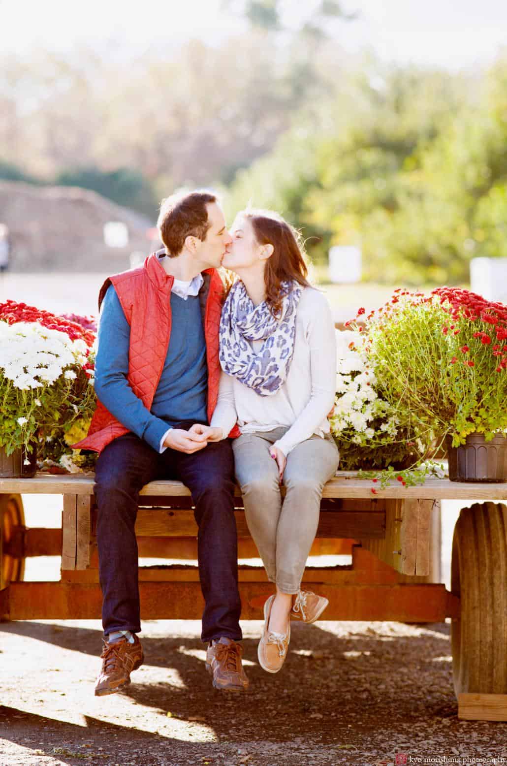 A couple sits on wooden crates surrounded by mums and kisses during their Terhune Orchards engagement shoot. One of NJ's best photoshoot locations, Terhune Orchards is located in Princeton.