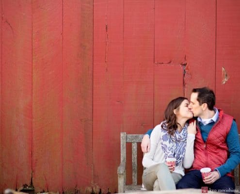 One of the best photography spots in NJ: the red barn at Terhune Orchards in Princeton. In this engagement picture, a couple hold cups of steaming hot apple cider and give each other a kiss.