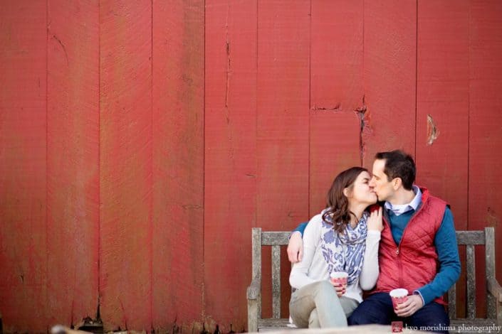 One of the best photography spots in NJ: the red barn at Terhune Orchards in Princeton. In this engagement picture, a couple hold cups of steaming hot apple cider and give each other a kiss.