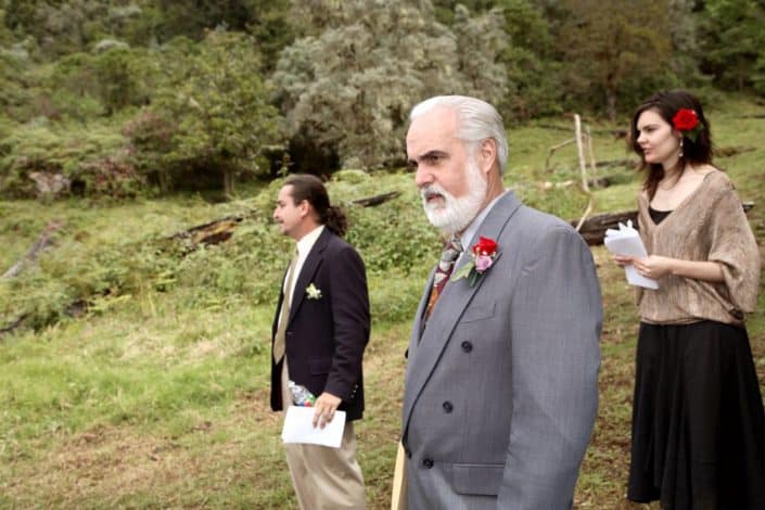 Father of the groom and two guests wait for bride on a mountaintop in San Gerardo de Dota for Costa Rican destination wedding. Girl with red rose in her hair, grey suit, red and pink rose boutonnnieres, Talamanca cloud forest background.