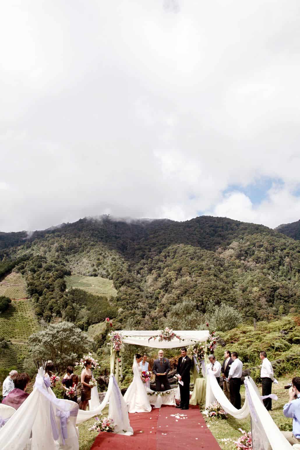 San Gerardo de Dota, Talamanca cloud forest wedding ceremony at Costa Rican destination wedding. Red carpeted aisle, white aisle swag with purple ribbons, bride, groom and wedding party surrounded by green, forest covered mountains.