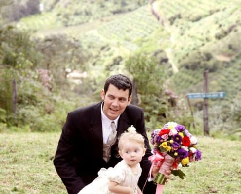 Groom and tiny flower girl on red carpet aisle runner at San Gerardo de Dota, Talamanca clound forest wedding ceremony. Purple, yellow and red bridal bouquet with pink ribbon, green mountaintop, Costa Rican destination wedding photographer.