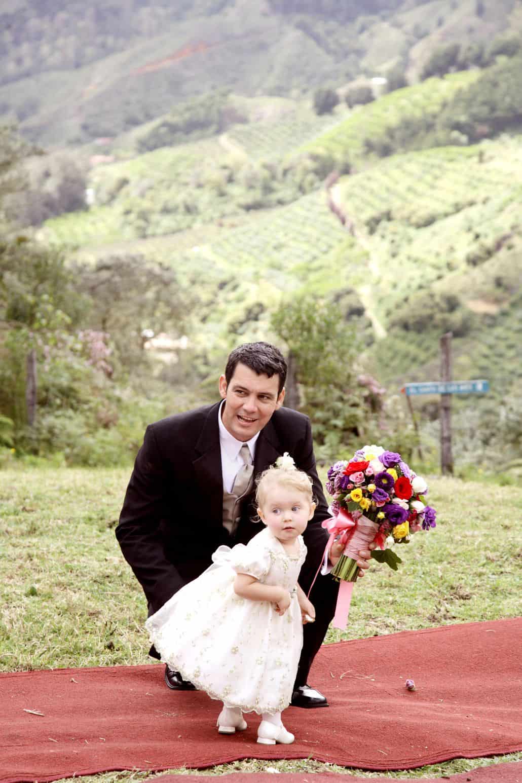 Groom and tiny flower girl on red carpet aisle runner at San Gerardo de Dota, Talamanca clound forest wedding ceremony. Purple, yellow and red bridal bouquet with pink ribbon, green mountaintop, Costa Rican destination wedding photographer.