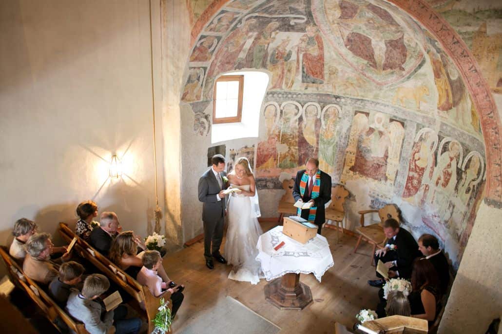 Bride and groom read together in tiny Romanesque church in Swiss Alps during European destination wedding ceremony. Domed alcove with painted mural.