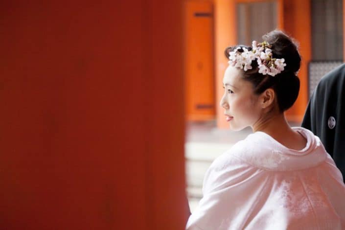 Bride looks over her shoulder at Heian Shrine in Kyoto, Japan. White kimono wedding dress and pink cherry blossoms in bride's hair. vivid orange woodwork. destination wedding photographer.