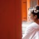 Bride looks over her shoulder at Heian Shrine in Kyoto, Japan. White kimono wedding dress and pink cherry blossoms in bride's hair. vivid orange woodwork. destination wedding photographer.