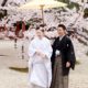 Bride smiles with guest under yellow paper umbrella on gravel path in front of blooming cherry blossom tree in Spring at destination wedding in Kyoto, Japan. Bride wears white kimono wedding dress and cherry blossoms in her hair.
