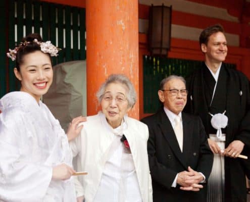 Bride and groom smile with grandparents, grandfather sticks his tongue out, at Heian Shrine in Kyoto, Japan for destination wedding photography. Bride wears white kimono and pink cherry blossoms in her hair, carries fan, groom wears montsuki and carried fan, large orange wooden column background.
