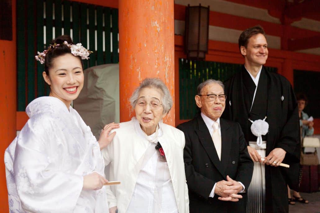 Bride and groom smile with grandparents, grandfather sticks his tongue out, at Heian Shrine in Kyoto, Japan for destination wedding photography. Bride wears white kimono and pink cherry blossoms in her hair, carries fan, groom wears montsuki and carried fan, large orange wooden column background.