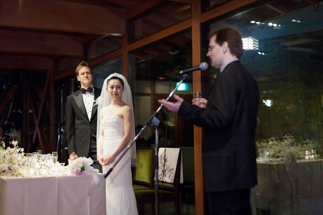 Guest gives speech at destination wedding in Kyoto, Japan, Garden Oriental, now called The Sodoh Higashiyama Kyoto, as bride and groom look on, Bride wears elegant white wedding dress with ruched bodice. floor to ceiling windows, pink and white bouquet.