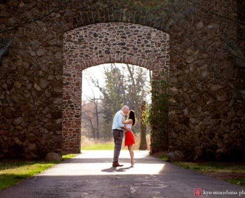 Couple kisses in front of arched, mossy, stone wall doorway for Hillsborough NJ photographer. Woman wears red skirt and white blouse. Man wears light blue gingham shirt and grey pants. French chateau, European estate feel.