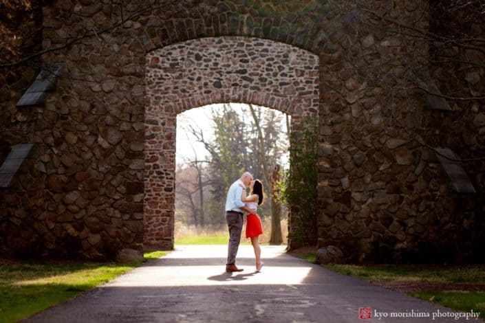 Couple kisses in front of arched, mossy, stone wall doorway for Hillsborough NJ photographer. Woman wears red skirt and white blouse. Man wears light blue gingham shirt and grey pants. French chateau, European estate feel.