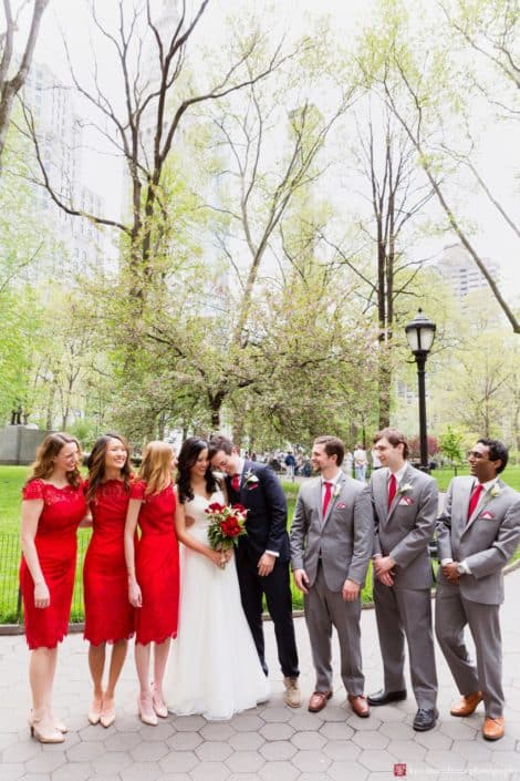 Groom leans head against bride surrounded by wedding party at madison square park wedding photo shoot NYC. red lace bridesmaid dresses from Lulu's, grey groomsmen suits with red ties, David's Bridal wedding dress, red and white wedding bouquet, Spring NYC wedding photos..