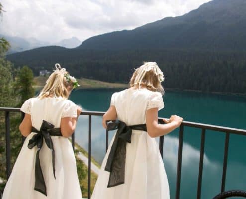 Flower girls in ivory dresses with black sashes lean on iron railing and overlook Lake St. Moritz at European destination wedding in Switzerland. Mountain and cloud background.