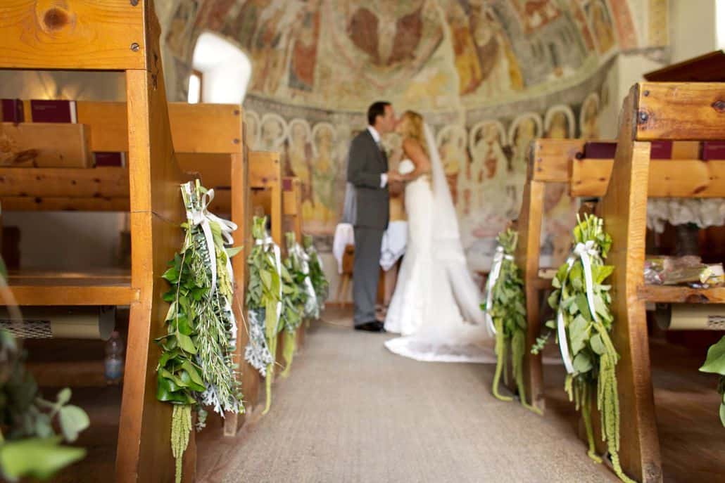 Bride and groom kiss at end of aisle in tiny Romanesque church at European destination wedding in Swiss Alps. Green and white bouquet pew arrangements, domed wall with aged, painted mural. Gardenias Floral.