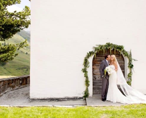 Groom kisses brides cheek in front of green floral covered archway on white wall at European destination wedding in Swiss Alps, destination wedding photography.