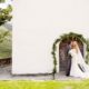 Groom kisses brides cheek in front of green floral covered archway on white wall at European destination wedding in Swiss Alps, destination wedding photography.