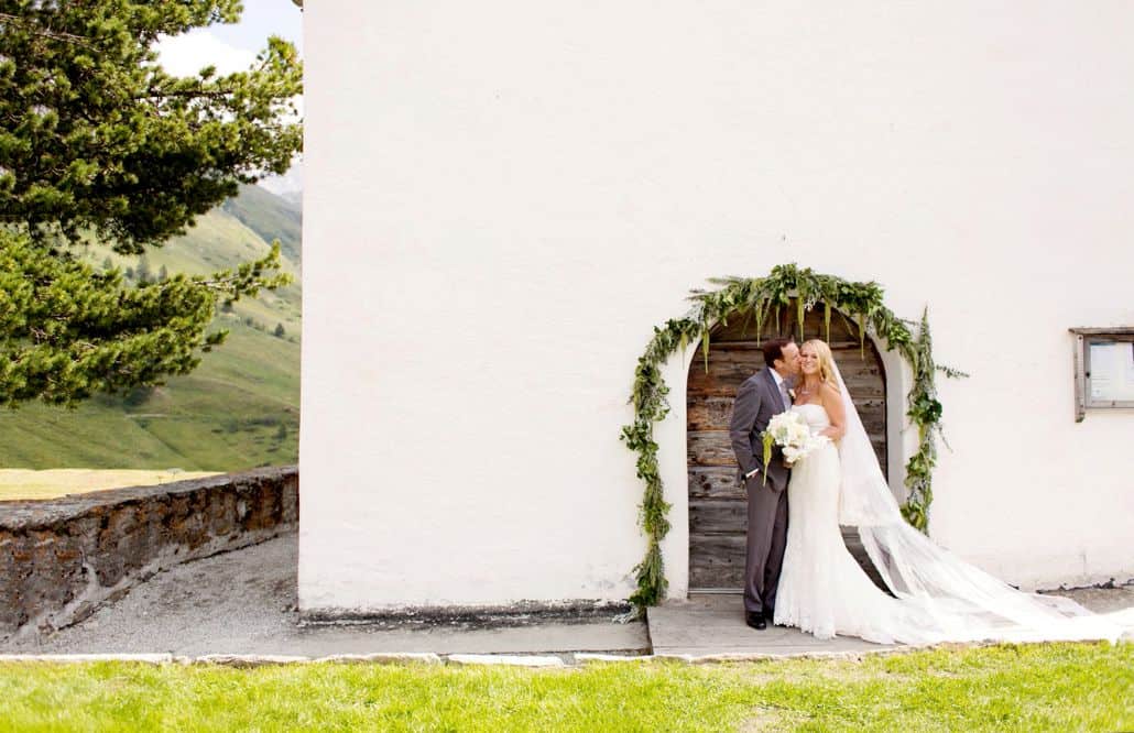 Groom kisses brides cheek in front of green floral covered archway on white wall at European destination wedding in Swiss Alps, destination wedding photography.