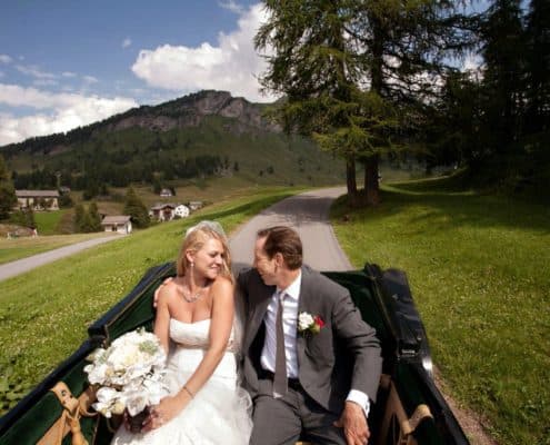 Bride and groom smile at each other in buggy ride at European destination wedding, white rustic wedding bouquet, strapless wedding dress with satin sash, Swiss Alps mountain background, winding road, small village.