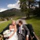 Bride and groom smile at each other in buggy ride at European destination wedding, white rustic wedding bouquet, strapless wedding dress with satin sash, Swiss Alps mountain background, winding road, small village.