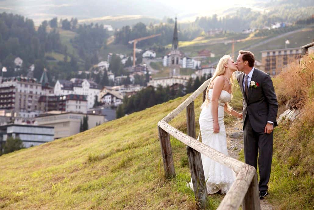 Bride and groom kiss next to old wooden fence on winding dirt path overlooking village in mountains, European destination wedding photography, Switzerland.