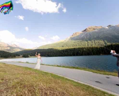 Bride and groom fly rainbow colored kite along path on Lake St. Moritz in Switzerland. Mountain and blue sky background. European destination wedding photographer,