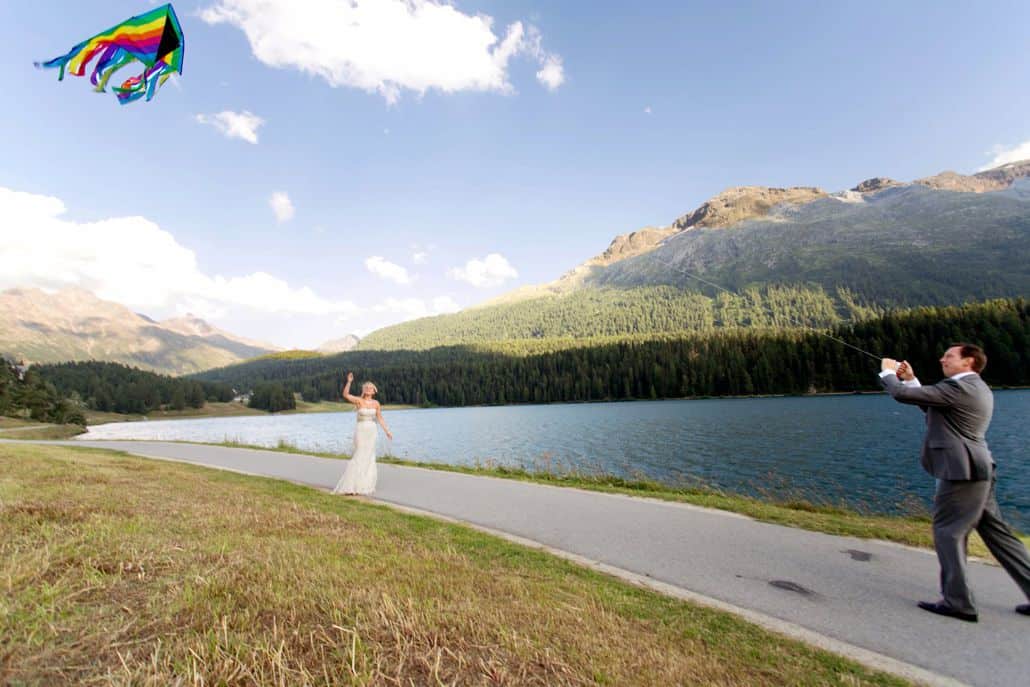 Bride and groom fly rainbow colored kite along path on Lake St. Moritz in Switzerland. Mountain and blue sky background. European destination wedding photographer,