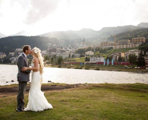 Bride and groom kiss in front of Lake St. Moritz in Switzerland, village along mountainside in background, white strapless wedding dress with satin sash, groom wears grey suit. European destination wedding photographer.