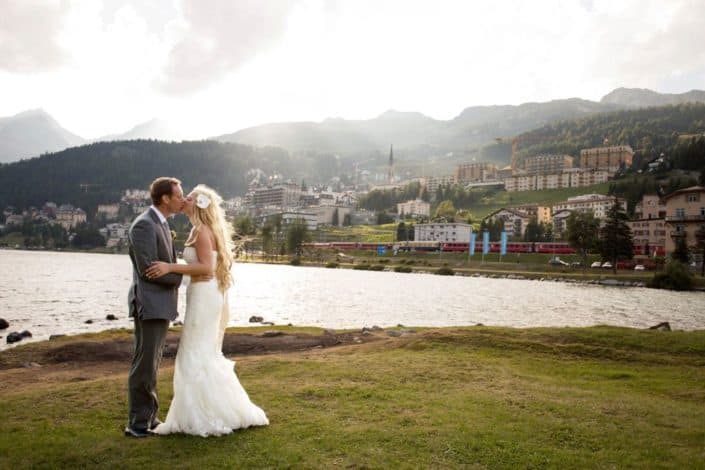 Bride and groom kiss in front of Lake St. Moritz in Switzerland, village along mountainside in background, white strapless wedding dress with satin sash, groom wears grey suit. European destination wedding photographer.