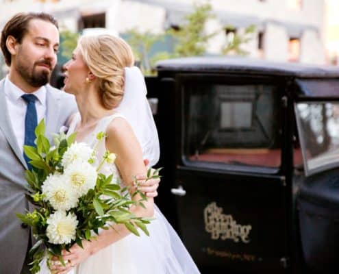 bride holds white dahlia and green foliage bridal bouquet while groom wraps his arm around her in front of vintage truck. groom wears grey suit and blue tie. flower wholesaler, flower district manhattan, the green building brooklyn NY, New York wedding photographer. DIY wedding flowers