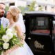 bride holds white dahlia and green foliage bridal bouquet while groom wraps his arm around her in front of vintage truck. groom wears grey suit and blue tie. flower wholesaler, flower district manhattan, the green building brooklyn NY, New York wedding photographer. DIY wedding flowers