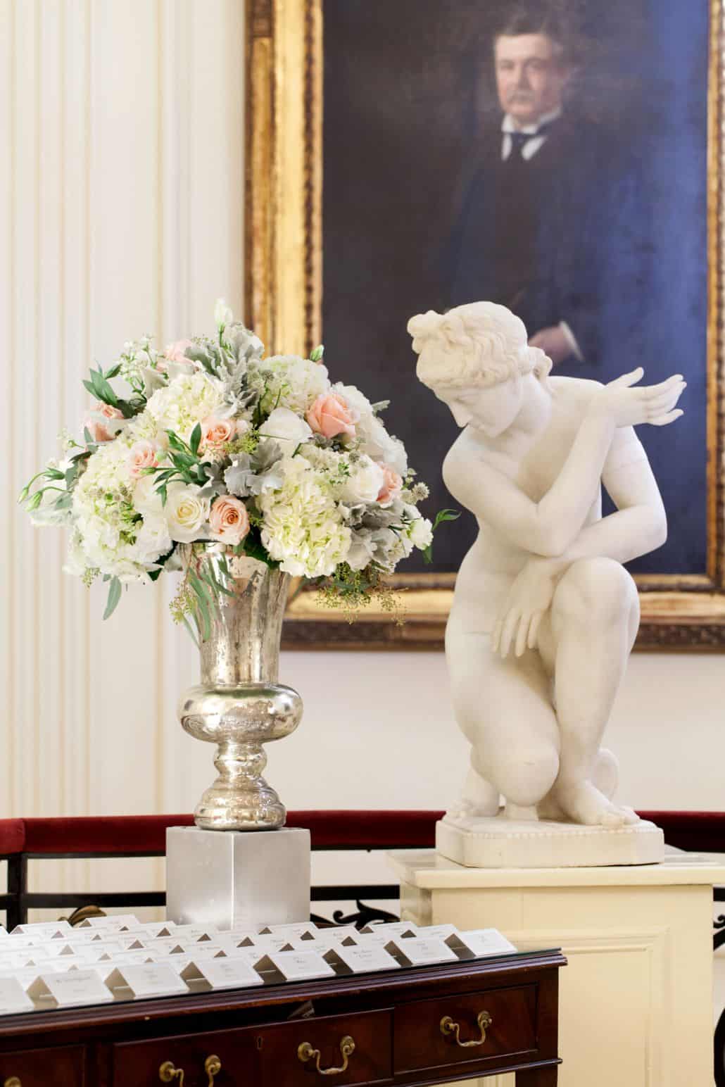 white peach and sage wedding floral arrangement at Lotos Club NYC, Fleur du Mois florist, antiqued silver vase next to statue on dark wood table with name cards. NYC wedding photographer.