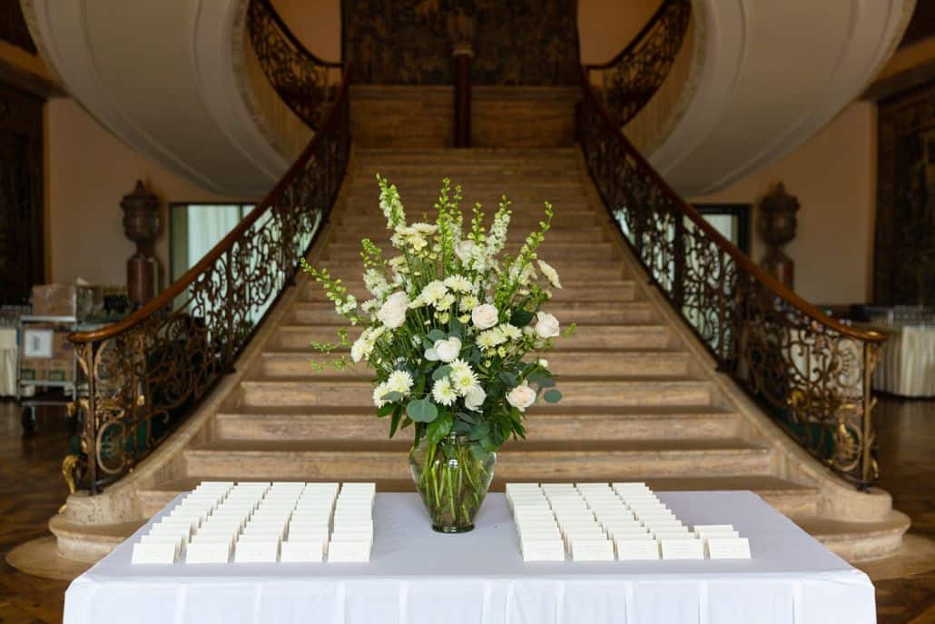 White and green floral arrangement in center of name card table at bottom of grand staircase at Jasna Polana, Princeton NJ wedding photographer, Janet Makrancy's Weddings and Parties Florist.