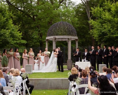 bride and groom kiss for first time as man and wife at non-denominational wedding ceremony at Jasna Polana under rounded top gazebo. Pronovias wedding gown. bridesmaids in greige Azazie dresses carrying baby's breath bouquets by Janet Mckrancy's wedding parties, groomsmen in Jos . A. Bank. suits. outdoor wedding ceremony. Jasna Polana wedding photographer.