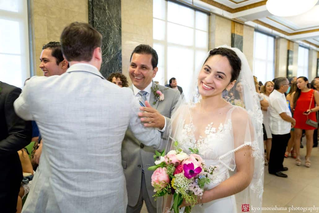 bride smiles at camera carrying pink, fuschia and white bouquet while father congratulates groom at NYC City Hall wedding. Homecoming florist. Elopement wedding photographer.